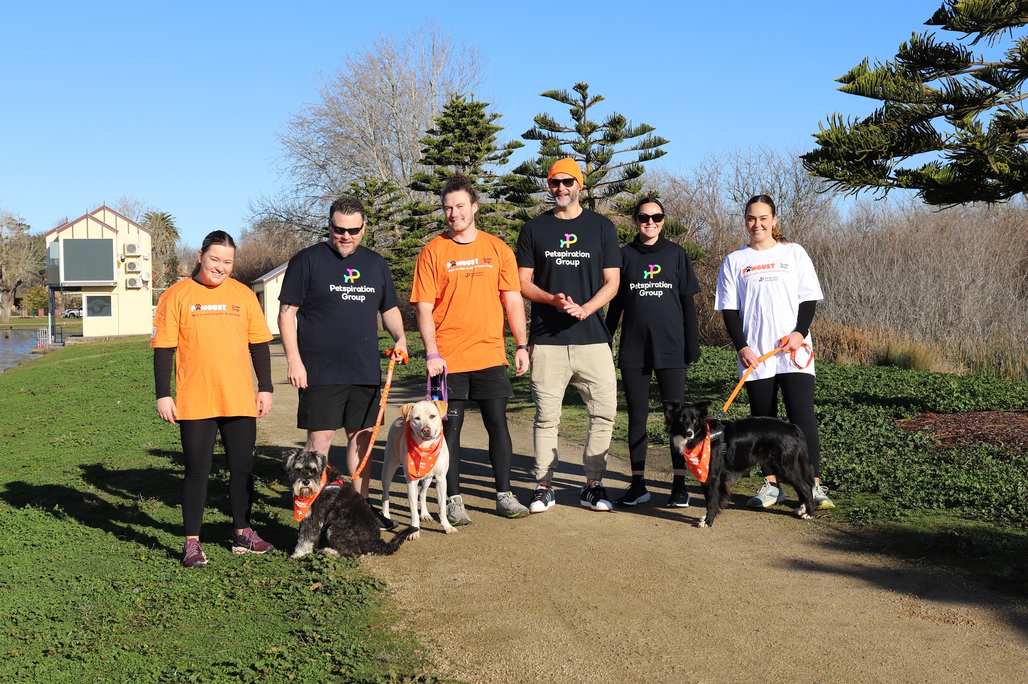 A group of 6 people standing on a footpath with 3 dogs on lead wearing orange and PAWGUST merch and Petspiration t-shirts.