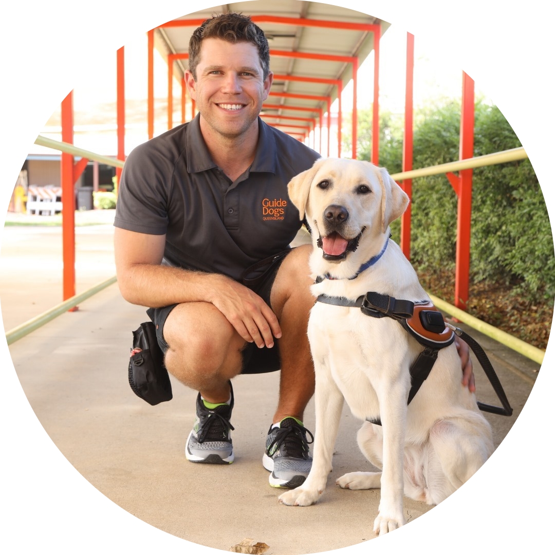 Image shows Guide Dog trainer sitting down next to a blonde Guide Dog wearing a harness. Image shows Guide Dog trainer sitting down next to a blonde Guide Dog wearing a harness.