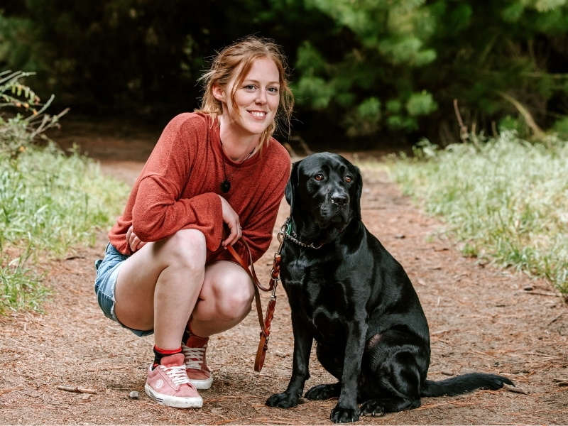A woman kneels next to her black guide dog in a forest.