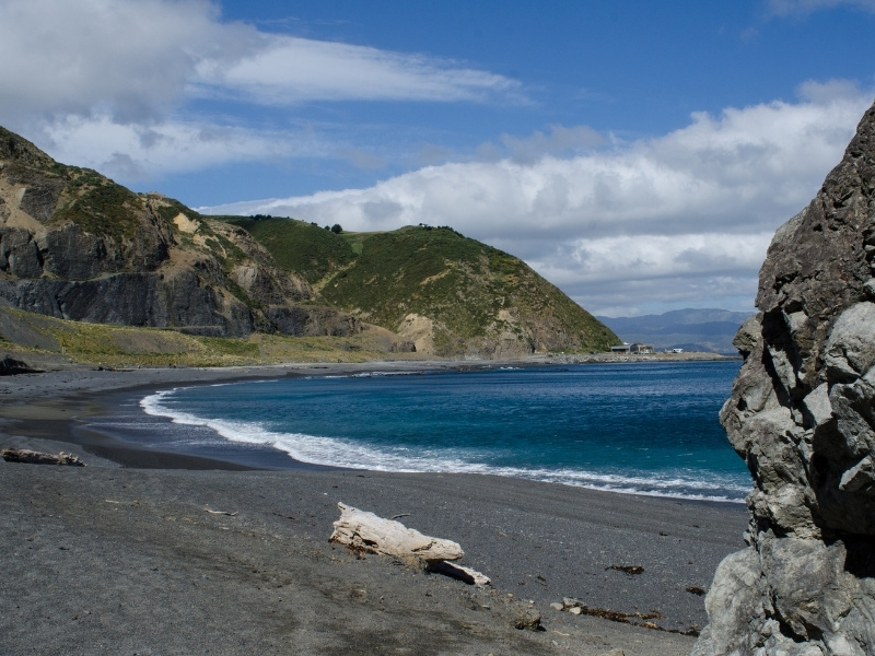 An image of a black sand beach, surrounded by mountains, on a sunny day