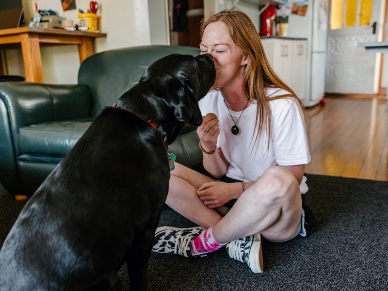 A woman sits cross legged in her lounge whilst her black guide dog boops her on the nose