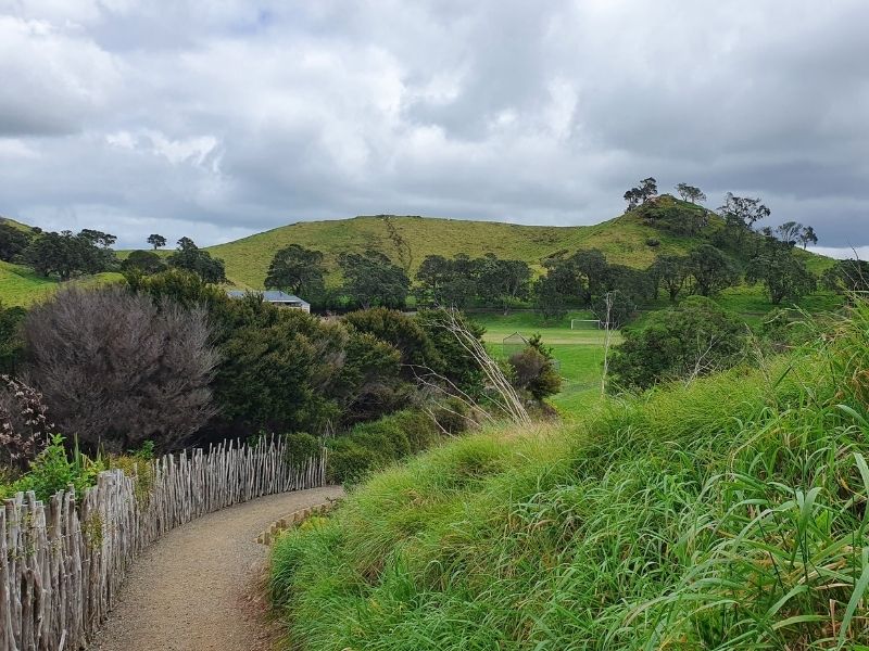 A path through a park. There is a grass bank on the right and a fence on the left. In the distance you can see a field.